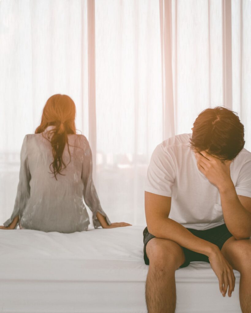 Couple sitting on opposite sides of the bed, man is looking down with head in hand