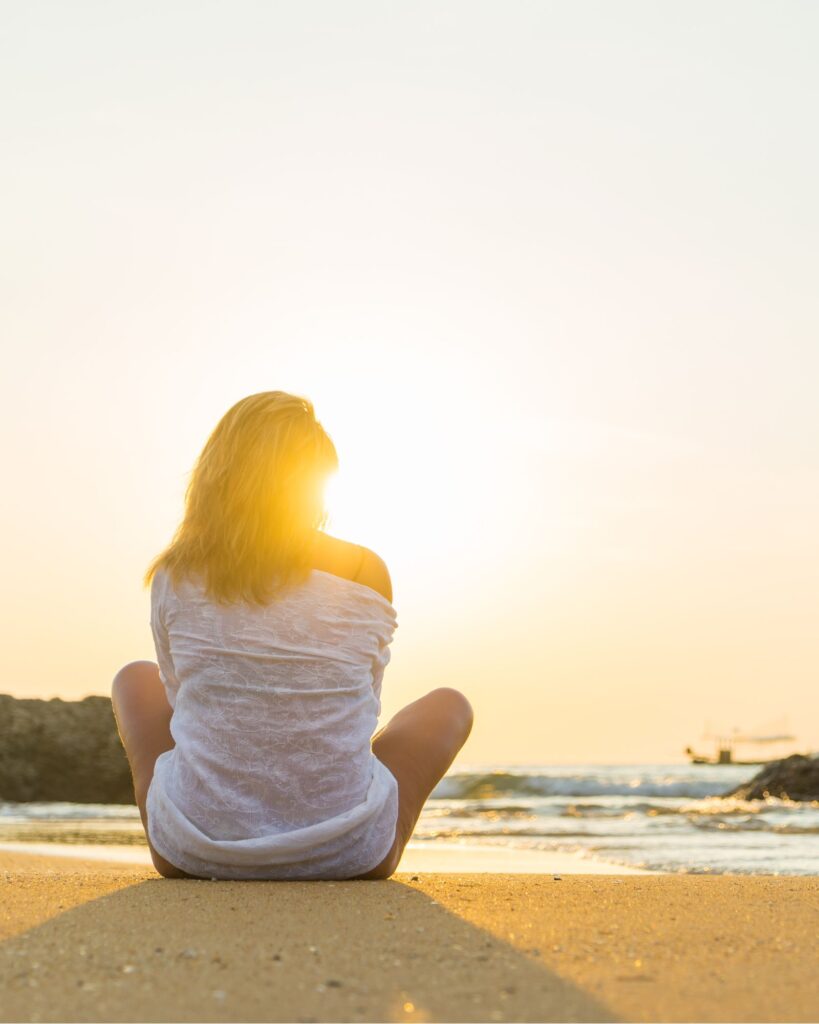Woman sitting crosslegged on a beach staring at the sunsest